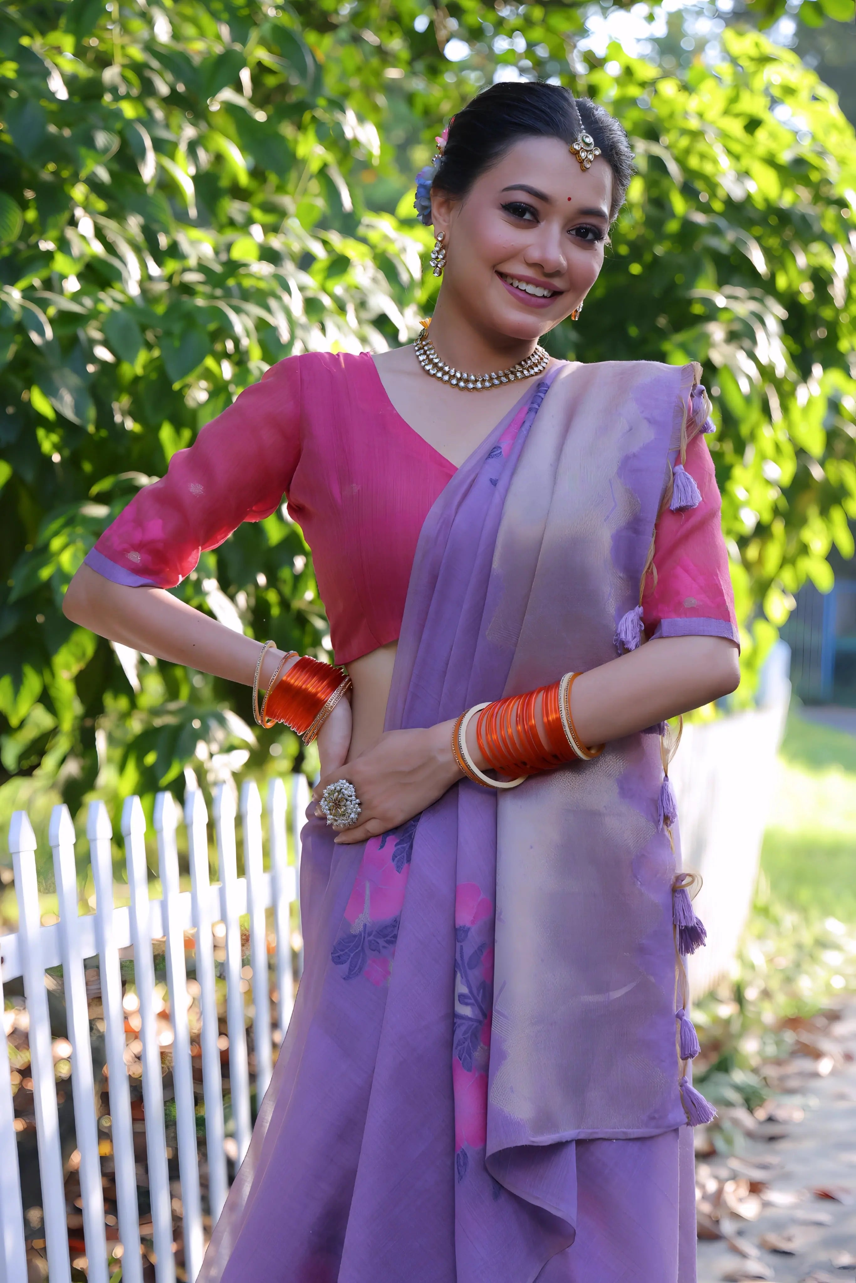 A woman models an elegant Lavender Purple Soft Muga Cotton Saree. The pallu features subtle Resham floral weaving in pink and purple. She wears a contrasting bright pink blouse and is accessorized with a pearl choker necklace and vibrant orange bangles, posing outdoors by a white fence.